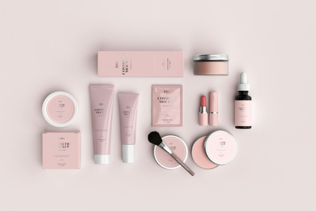 Pink cosmetic tubes, compacts and a brush arranged on a pale background